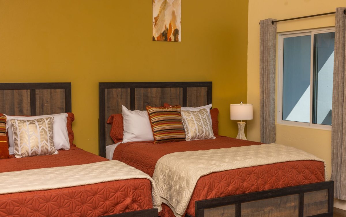 Two beds in a room with orange bedding and decorative pillows, next to a window with gray curtains.