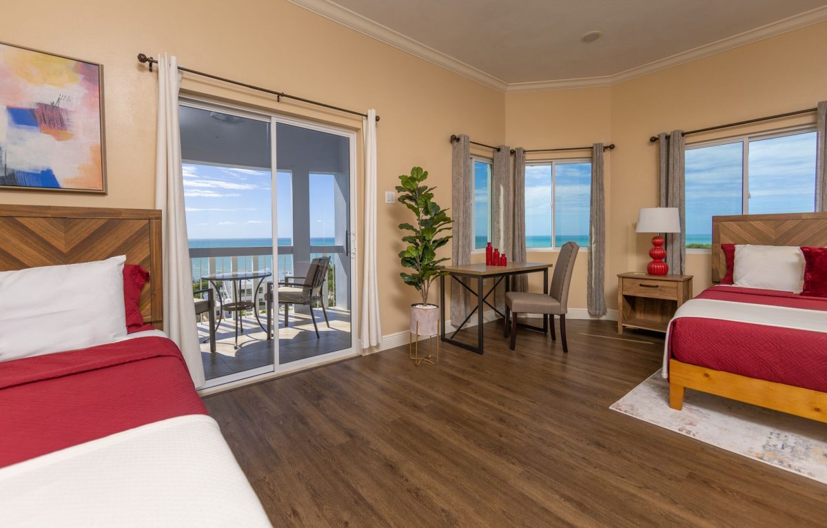 Bedroom with two beds, balcony overlooking the ocean, desk by window, warm tones.
