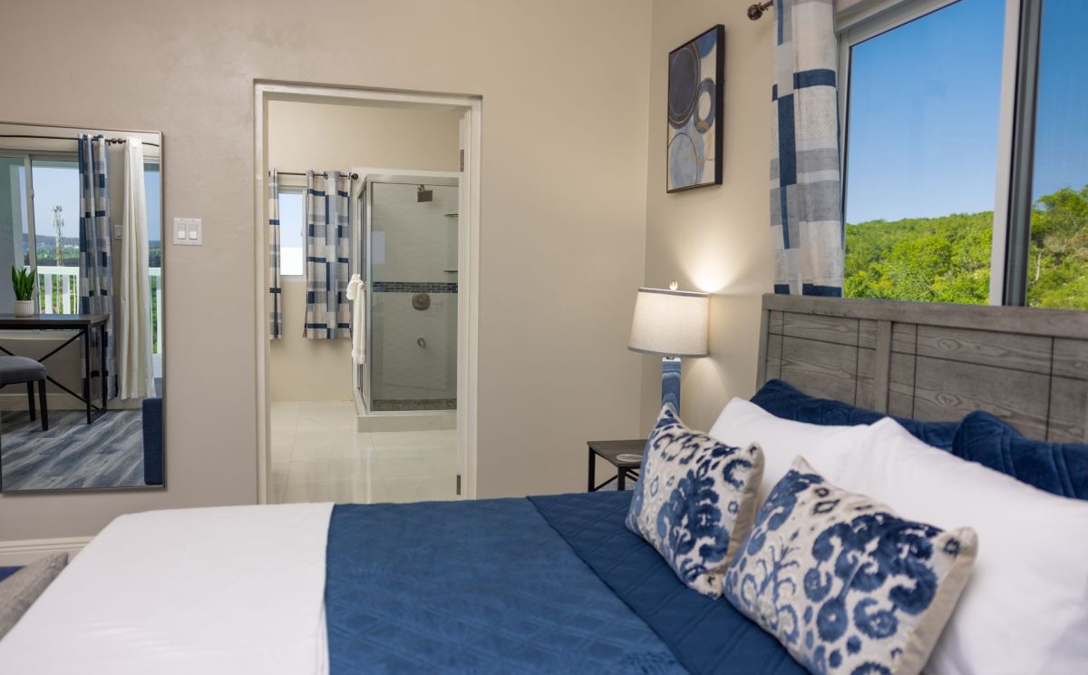 Bedroom with blue and white bedding, wooden headboard, open door to bathroom, and window with trees.